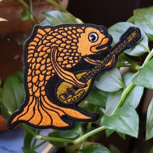 May include: A yellow and black embroidered patch of a fish playing a guitar. The fish has a detailed design with scales and fins. The guitar is black with white strings.