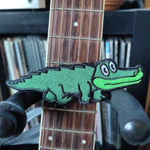 May include: A green embroidered alligator patch with white eyes is attached to a guitar neck. The guitar neck is brown with white fret markers. The patch is on a black guitar stand.