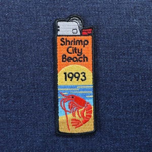 Può includere: Toppa ricamata a forma di accendino, con la scritta "Shrimp City Beach 1993". Il design include un'illustrazione di gambero rosso su una scena di spiaggia con acqua blu e sabbia gialla.
