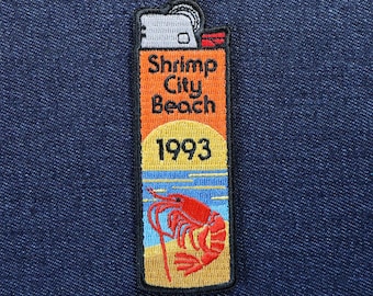 May include: Embroidered patch in the shape of a lighter, featuring the text "Shrimp City Beach 1993." The design includes a red shrimp illustration on a beach scene with blue water and yellow sand.