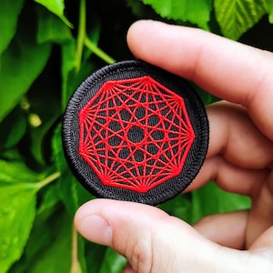 May include: A circular embroidered patch featuring a black fabric base with a vibrant red geometric pattern. The design consists of intricate, intersecting lines forming a complex star-like shape within a circular border, all rendered in red thread.