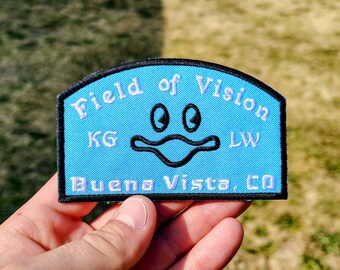 May include: A light blue embroidered patch with a black border, featuring the text Field of Vision at the top, Buena Vista, CO at the bottom, and the letters KG and LW on the sides. A central black line drawing depicts a stylized duck face.