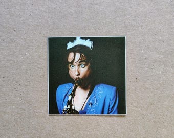 May include: A square sticker featuring a person with wide eyes wearing a white headpiece and a bright blue jacket with embroidered details, playing a saxophone against a dark background. The image captures a focused expression while performing.