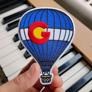 May include: An embroidered patch shaped like a hot air balloon with a blue and white striped pattern. A large red letter C with a yellow center is stitched onto the balloon. The patch features a small brown basket at the bottom with black stitching.