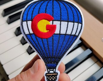 May include: An embroidered patch shaped like a hot air balloon with a blue and white striped pattern. A large red letter C with a yellow center is stitched onto the balloon. The patch features a small brown basket at the bottom with black stitching.