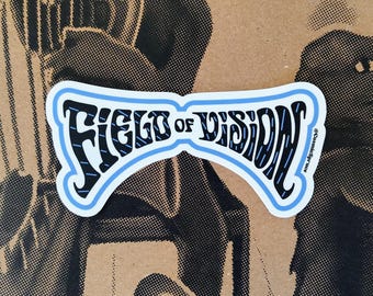 May include: A die-cut sticker featuring the text FIELD OF VISION in a stylized black font with blue vertical line accents. The lettering is enclosed within a light blue border that follows the curved shape of the words. The text CosmicSpruce is printed in small black letters on the right side of the sticker.
