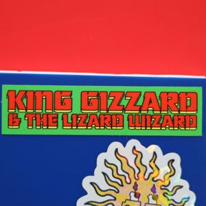 Könnte beinhalten: Ein grüner und gelber Aufkleber mit dem Text "KING GIZZARD & THE LIZARD WIZARD" in roten und gelben Buchstaben. Der Aufkleber befindet sich auf einer blauen Oberfläche.