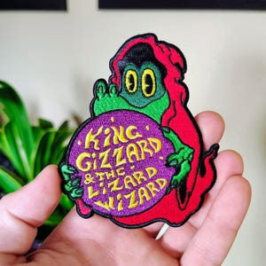 Könnte beinhalten: Ein gestickter Aufnäher, der eine grüne Kreatur mit roter Kapuze zeigt, die eine violette Kugel hält. Auf der Kugel steht in gelber Schrift "KING GIZZARD & THE LIZARD WIZARD". Der Aufnäher hat einen schwarzen Rand und detaillierte Stickereien.