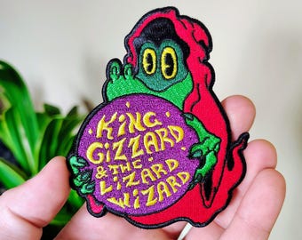 May include: An embroidered patch featuring a green creature with a red hood holding a purple orb. The orb displays the yellow text "KING GIZZARD & THE LIZARD WIZARD" in a stylized font. The patch has a black border and detailed stitching throughout.