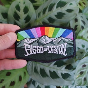 Peut inclure: Un écusson brodé rectangulaire à bordure noire représentant une chaîne de montagnes dans des tons de gris et de vert sous un soleil arc-en-ciel vibrant. Le texte "FIELD OF VISION" est brodé en lettres blanches stylisées sur la partie inférieure.