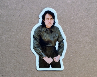 May include: A die-cut sticker featuring a person with dark hair and a mustache, wearing a dark olive green long-sleeved jumpsuit with a front zipper and chest pockets. The person has a serious expression and is clenching both fists near their waist.