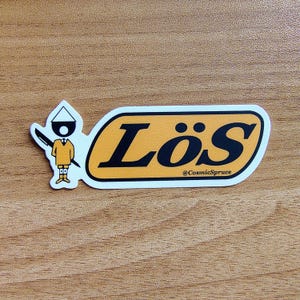 May include: A sticker with the word "Lös" in black letters on an orange background, with a white border. A cartoon figure with a pen is on the left. The text "@CosmicSpruce" is below the word "Lös".