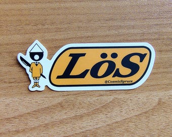May include: A sticker with the word "Lös" in black letters on an orange background, with a white border. A cartoon figure with a pen is on the left. The text "@CosmicSpruce" is below the word "Lös".