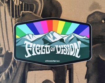 May include: A rectangular sticker with rounded corners featuring a mountain range in shades of blue and white against a vibrant rainbow sunburst background. The text FIELD OF VISION is printed in white, with the handle @CosmicSpruce below it.