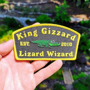May include: A black embroidered patch with a yellow border featuring the text King Gizzard and Lizard Wizard in yellow thread. A green cartoon crocodile is centered between the text with the letters EST. and the number 2010 on either side.