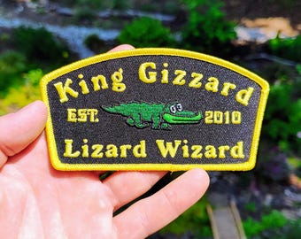 May include: A black embroidered patch with a yellow border featuring the text King Gizzard and Lizard Wizard in yellow thread. A green cartoon crocodile is centered between the text with the letters EST. and the number 2010 on either side.