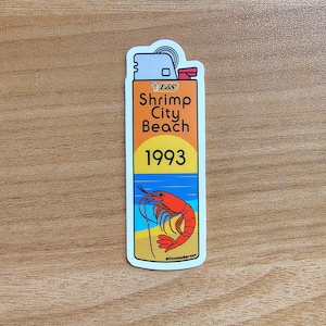 May include: A sticker in the shape of a lighter with the text "Shrimp City Beach 1993." The design features a shrimp, beach, and ocean scene in orange, yellow, blue, and white. The sticker is on a wooden surface.
