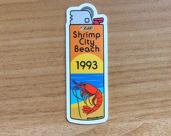 May include: A sticker in the shape of a lighter with the text "Shrimp City Beach 1993." The design features a shrimp, beach, and ocean scene in orange, yellow, blue, and white. The sticker is on a wooden surface.