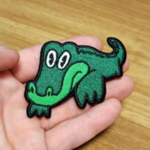 Op de afbeelding: Geborduurde groene alligator patch met een cartoon ontwerp. De patch heeft een groen lichaam, een lichtgroene tong en witte ogen met zwarte pupillen. De patch heeft een zwarte rand en is waarschijnlijk voor kleding of accessoires.