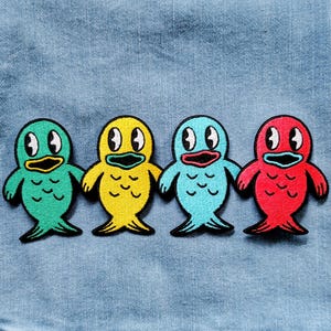 Puede incluir: Cuatro parches bordados con forma de pez en verde, amarillo, azul y rojo. Cada pez tiene contornos negros, ojos blancos y boca abierta. Los parches están sobre un fondo de tela vaquera azul.