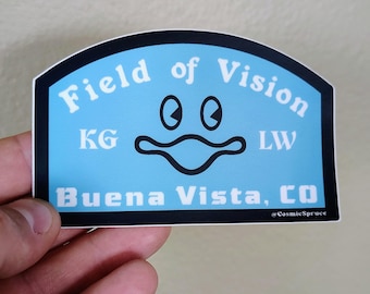 May include: A light blue sticker with a black border featuring the text Field of Vision, KG, LW, and Buena Vista, CO in white. A central black graphic depicts two oval eyes above a wide, wavy mouth shape. The text @CosmicSpruce is in the corner.