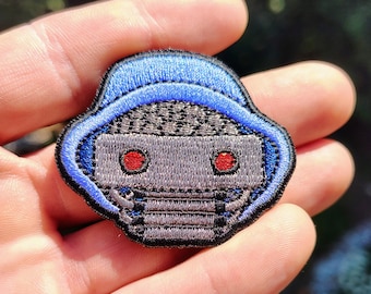 May include: An embroidered patch featuring a stylized robot head with a grey face, two circular red eyes, and a textured grey top section. The robot is framed by a bright blue hood-like border, all outlined with a thick black stitched edge.