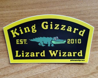May include: A rectangular sticker with a yellow border and black background featuring a green cartoon alligator in the center. The text reads King Gizzard EST. 2010 Lizard Wizard in yellow, with the small text @CosmicSpruce in the bottom right corner.