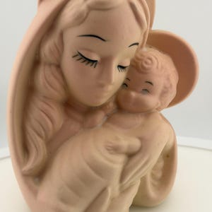 Puede incluir: Una figura de cerámica de color melocotón mate que representa una silueta estilizada sosteniendo a un niño pequeño. Ambas figuras tienen los ojos cerrados, pestañas definidas y rasgos suaves y redondeados. La figura mayor lleva un tocado.