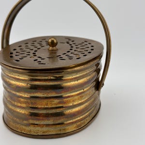Puede incluir: Un pequeño recipiente de latón de forma ovalada con asa y tapa perforada. El recipiente tiene un diseño acanalado y un acabado ligeramente deslustrado.