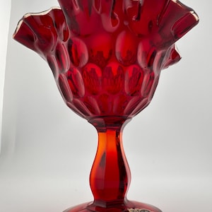 May include: A red glass candy dish with a ruffled rim and a textured, bubbled design. The dish has a tall, slender stem and a round base. The dish has a sticker on the bottom that says "Fiesta"