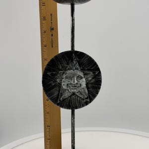 May include: A black metal candle holder with a distressed silver finish, featuring a circular disc with a raised star and sun face design. A wooden ruler shows the height is approximately 12 inches and the base width is about 6 inches.
