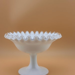 Puede incluir: Un cuenco de pedestal de vidrio blanco con borde ondulado. El cuenco tiene un borde ancho y festoneado y se apoya sobre un tallo y una base cortos y redondeados. El vidrio tiene un aspecto lechoso y opaco. Probablemente se utiliza para servir postres o exhibir objetos decorativos.
