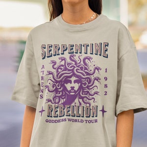 May include: A light beige t-shirt with a purple and black graphic of a woman with snakes for hair. The text "Serpentine Rebellion" is printed in a vintage style font with the words "Athens 1982" and "Goddess World Tour" printed below the graphic.