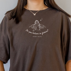 May include: An oversized brown t-shirt featuring a white graphic of a fairy sitting on a mushroom with the text "Do you believe in fairies?" and "J.M. Barrie - Peter Pan" underneath.
