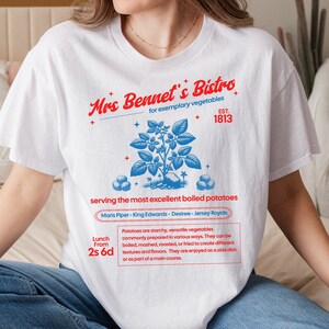 Op de afbeelding: Wit T-shirt met een grafisch ontwerp van een aardappelplant en de tekst "Mrs Bennet's Bistro for exemplary vegetables EST. 1813 serving the most excellent boiled potatoes Maris Piper - King Edwards - Desiree - Jersey Royals Lunch From 2s 6d Potatoes are starchy, versatile vegetables commonly prepared in various ways. They can be boiled, mashed, roasted, or fried to create different textures and flavours. They are enjoyed as a side dish or as part of a main course."