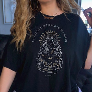May include: Black t-shirt with a gold graphic of a woman with a crown of stars and the text "I spoke to you Aphrodite, in a dream" and "Sappho" at the bottom.