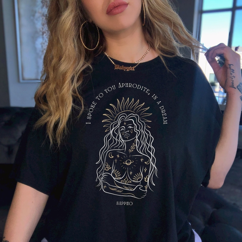 May include: Black t-shirt with a gold graphic of a woman with a crown of stars and the text "I spoke to you Aphrodite, in a dream" and "Sappho" at the bottom.