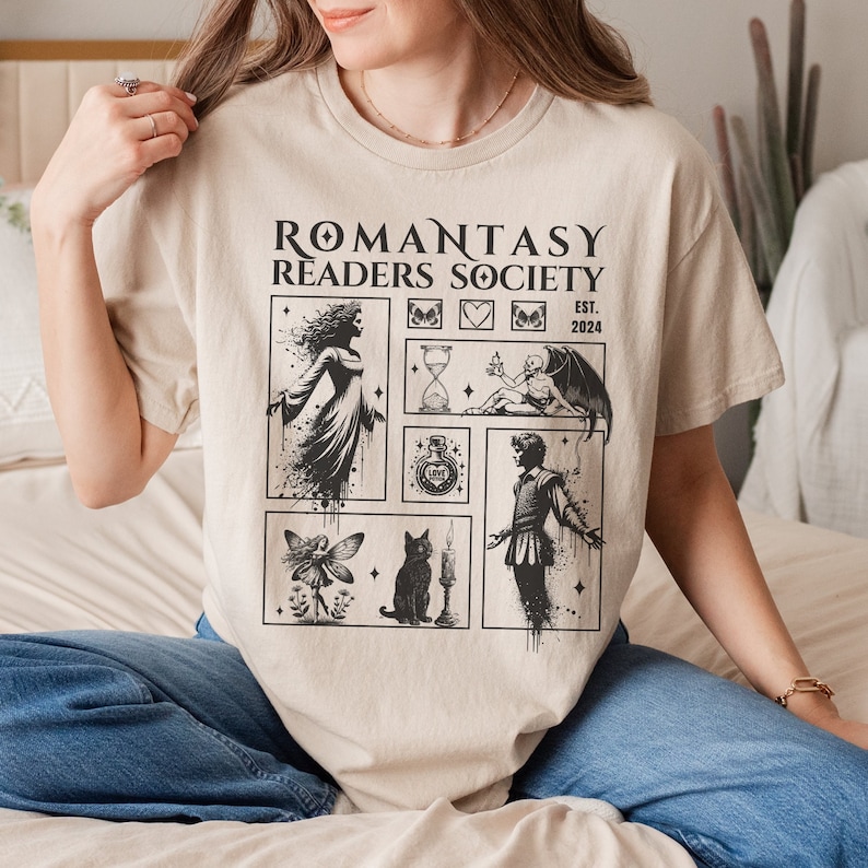 May include: A beige t-shirt with a black graphic print that says "Romantasy Readers Society Est. 2024". The graphic includes illustrations of a fairy, a cat, a potion bottle, a hourglass, a candle, a woman, a man, and a demon.