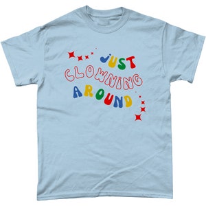Op de afbeelding: Lichtblauw T-shirt met de tekst "JUST CLOWNING AROUND" in kleurrijke, speelse letters.