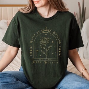 Może przedstawiać: Zielony T-shirt z nadrukiem w kolorze złotym przedstawiającym r&oacute;żę, koronę i tekst "Queen of England 1501 - 1536 Anne Boleyn".