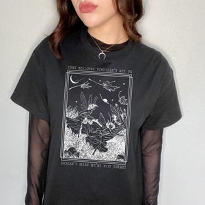 K&ouml;nnte beinhalten: Schwarzes T-Shirt mit einem wei&szlig;en Grafikdruck von Feen, die in einem Nachthimmel fliegen. Die Grafik enth&auml;lt den Text "Just because you can't see us doesn't mean we're not there!"