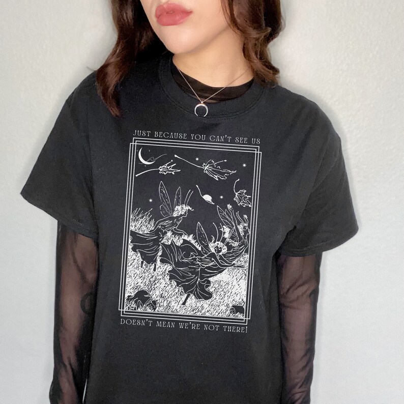 K&ouml;nnte beinhalten: Schwarzes T-Shirt mit einem wei&szlig;en Grafikdruck von Feen, die in einem Nachthimmel fliegen. Die Grafik enth&auml;lt den Text "Just because you can't see us doesn't mean we're not there!"