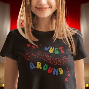 Op de afbeelding: Een zwart T-shirt met de tekst "Just Clowning Around" in kleurrijke, omrande letters. De letters zijn in een speels, afgerond lettertype.