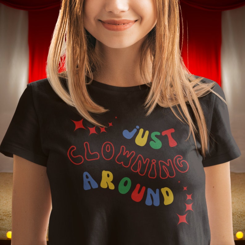 Op de afbeelding: Een zwart T-shirt met de tekst "Just Clowning Around" in kleurrijke, omrande letters. De letters zijn in een speels, afgerond lettertype.