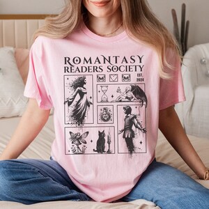 May include: Pink t-shirt with a black graphic print that says "Romantasy Readers Society Est. 2024" and features illustrations of a fairy, a cat, a potion bottle, an hourglass, a skeleton holding a candle, and a person in a cloak.