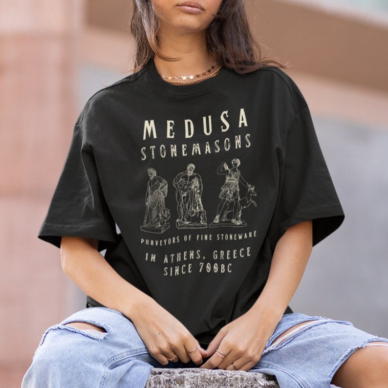 May include: Black t-shirt with a white graphic design that says "Medusa Stonemasons" and features three figures. The text below the figures says "Purveyors of Fine Stoneware in Athens, Greece since 700BC".
