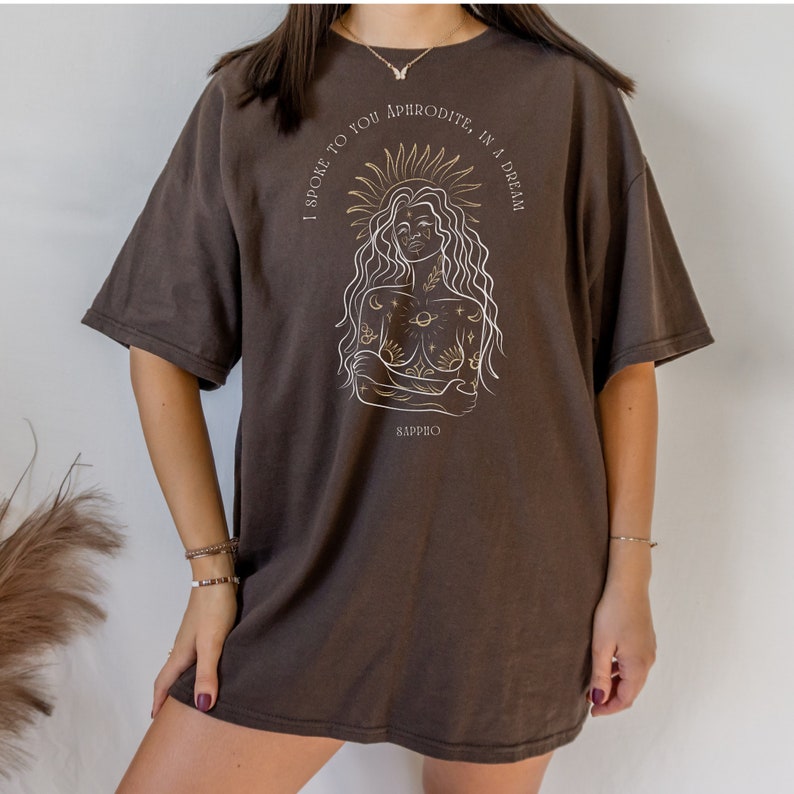 May include: A brown oversized t-shirt with a gold graphic of a woman with a sun halo and the text "I spoke to you Aphrodite in a dream Sappho".