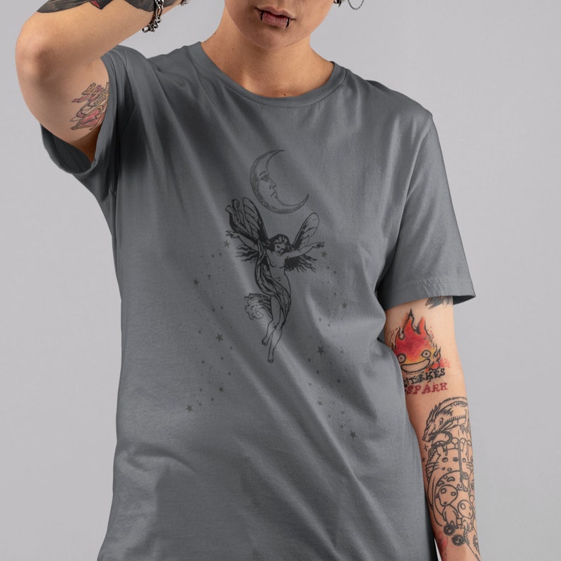May include: A dark gray t-shirt with a black and white graphic of a fairy flying with a crescent moon above her. The graphic is surrounded by small stars.