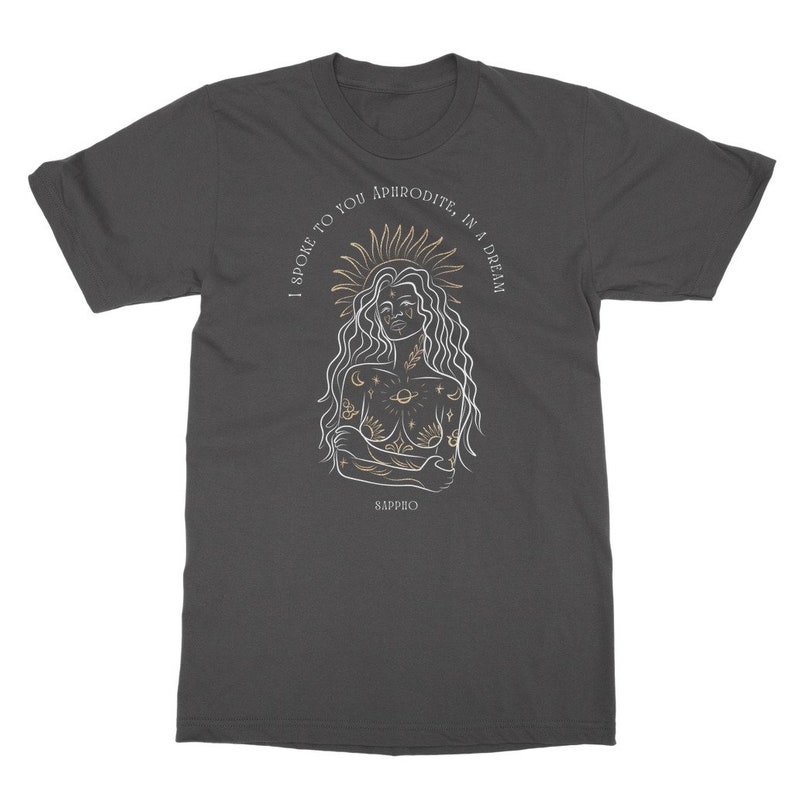 May include: A dark gray t-shirt with a white outline of a woman with celestial tattoos and the text "I spoke to you Aphrodite, in a dream" in gold lettering. The text "Sappho" is also in gold lettering at the bottom of the design.