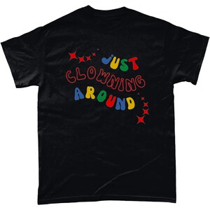 Op de afbeelding: Zwart T-shirt met de tekst "JUST CLOWNING AROUND" in kleurrijke, speelse letters. De tekst is omringd door kleine rode, gele en blauwe sterren.